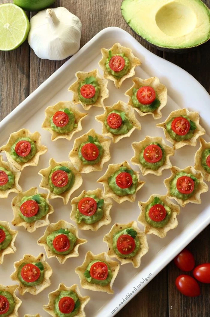 christmas party snacks, small taco shells filled with guacamole, halved cherry tomatoes on top, arranged on white tray