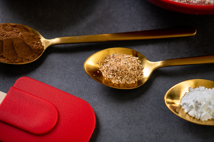 three brass spoons with different ingredients, red silicone spatula, placed on black surface, decorating gingerbread cookies