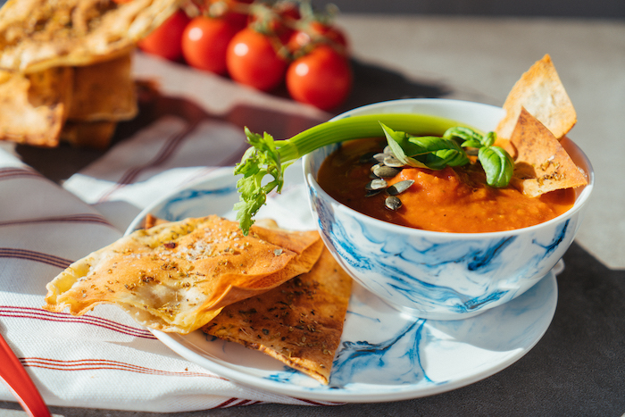 tomato soup recipe, tomato soup inside a ceramic bowl, garnished with seeds celery and basil, bread on the side