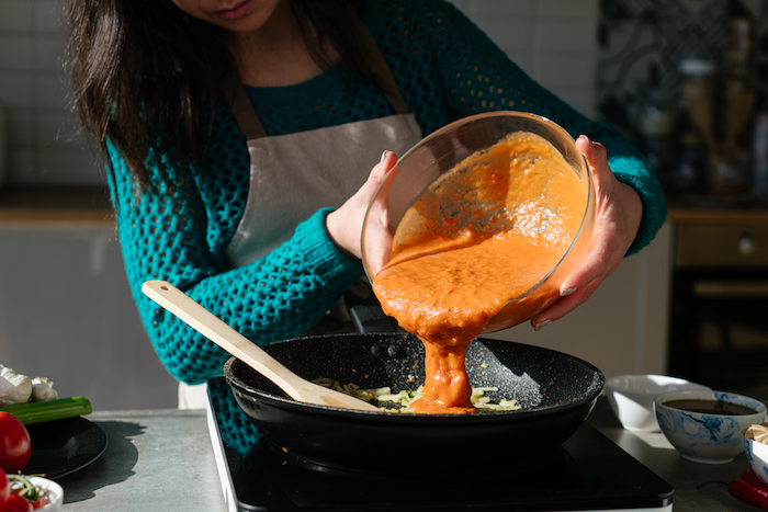 pureed tomatoes in glass bowl, tomato soup recipe, poured over celery and onion in black sauce pan, wooden spatula on the side