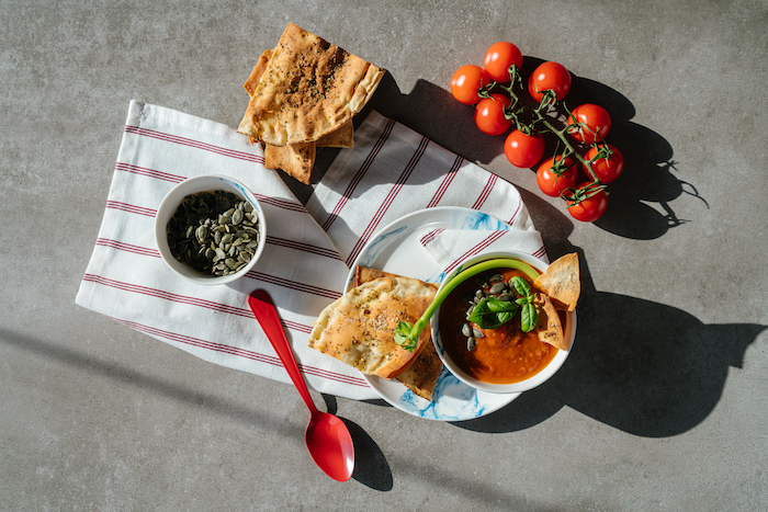 tomato soup, ceramic bowl full of soup, tomatoes and seeds, arranged on grey countertop, red and white cloth underneath