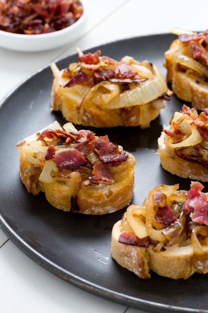 bruschetta with bacon and caramelised onion on top, christmas party snacks, arranged on black plate, placed on white wooden surface