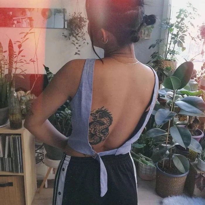 woman wearing black pants, open back denim top, dragon thigh tattoo, back tattoo, black hair, lots of plants in the background
