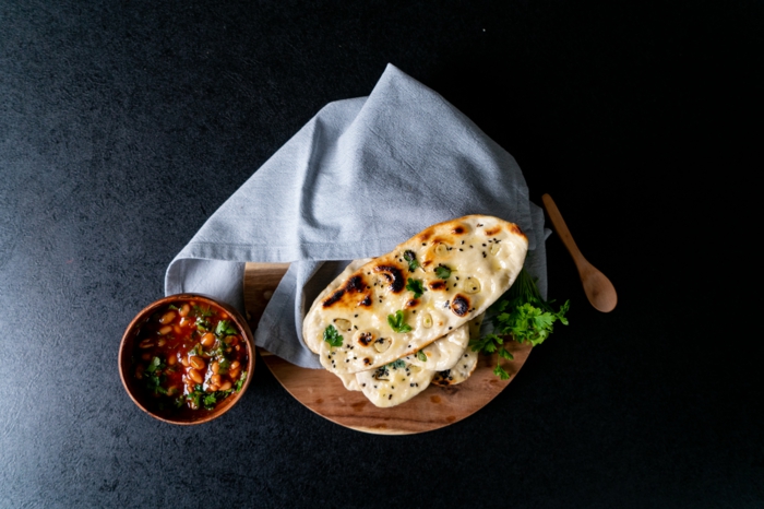 naan bread, placed on wooden cutting board, blue cloth on top of it, beans in tomato sauce in wooden bowl