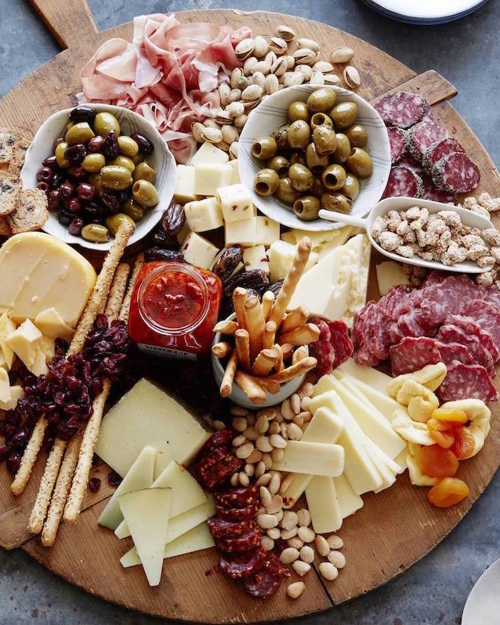 large round wooden board, cheese and meat on it, bowls of olives, christmas party snacks, crackers and pretzels