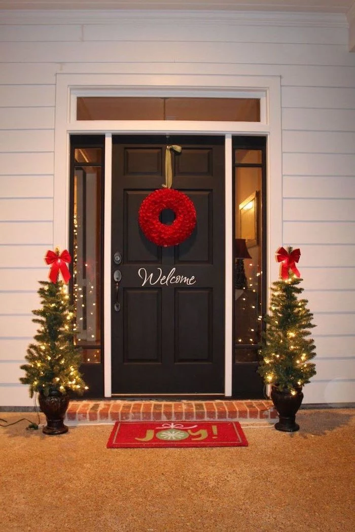 red wreath hanging on the door, outdoor christmas tree lights, two small trees with lights and red ribbons, on both sides of the door