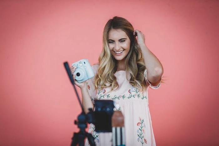 woman holding an instant camera, wearing pink floral dress, brunette highlights, long wavy brown hair with blonde balayage