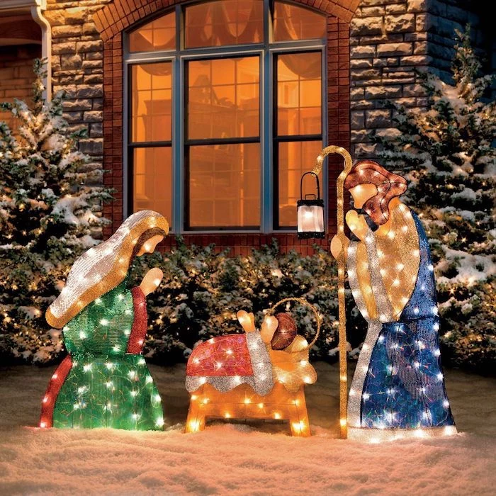 nativity scene with lights, christmas deer decorations, placed in the snow, in front of a house, bushes and trees decorated with lights