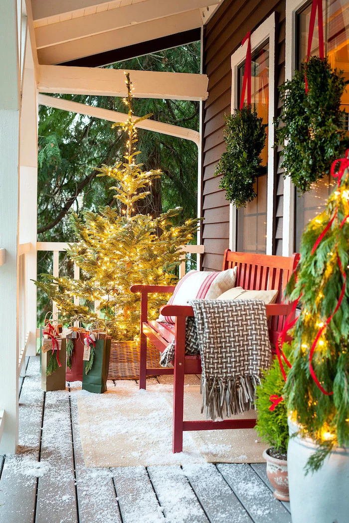 tree decorated with lights, placed on the front porch, next to a wooden bench, large outdoor christmas decorations