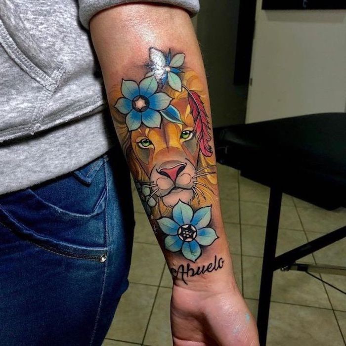 colored forearm tattoo, lion thigh tattoo, lion head surrounded by blue flowers, on woman wearing jeans and grey blouse