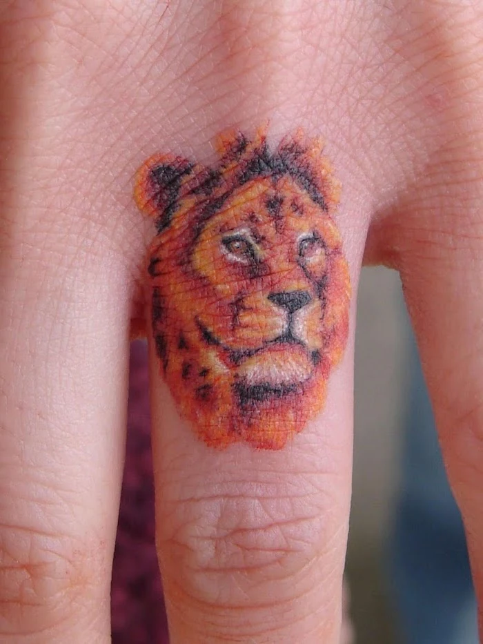 finger tattoo on the ring finger, small lion tattoo, lion head in orange and black