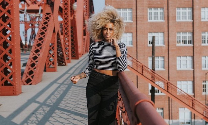 woman standing on a bridge, wearing black pants and striped blouse, winter hair colors, very curly blonde hair with dark roots