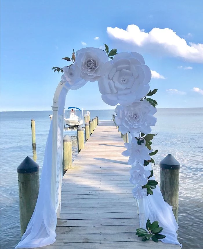 wooden arch, decorated with white tulle, large white paper roses, placed on a wooden pier, paper flower templates