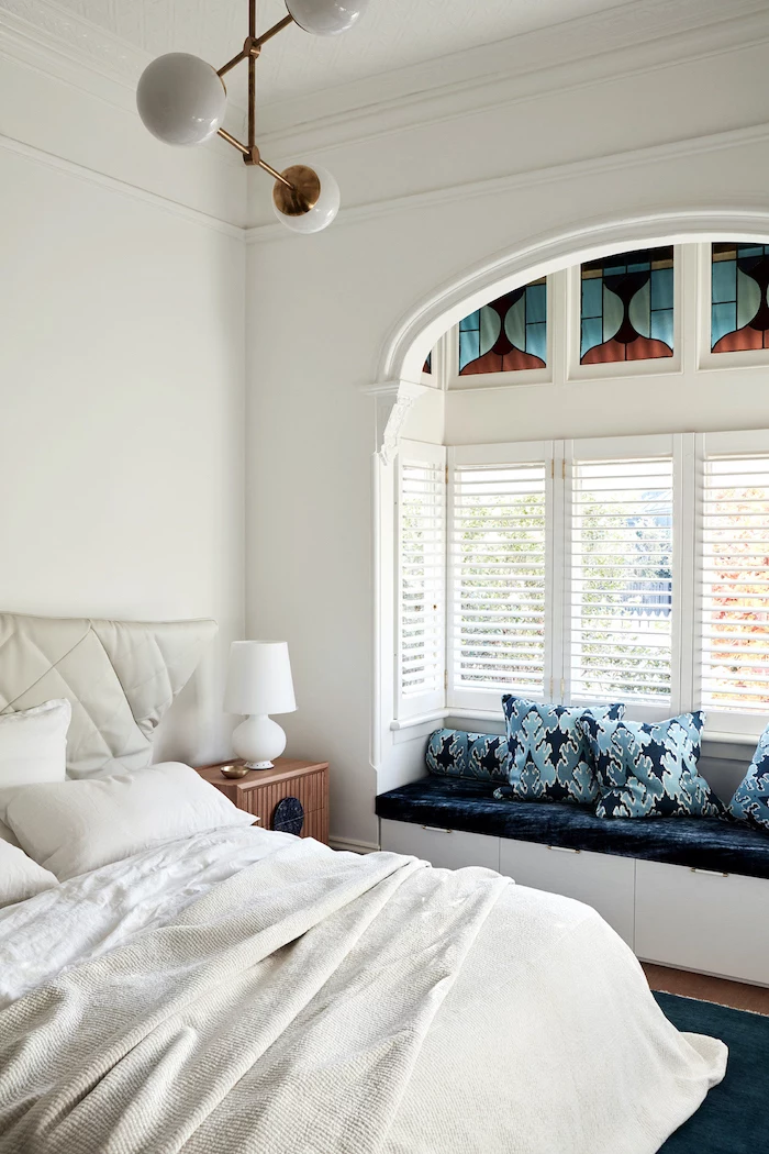 bedroom with white walls, large windows with blinds, stained glass panels, bench with blue velvet pillows