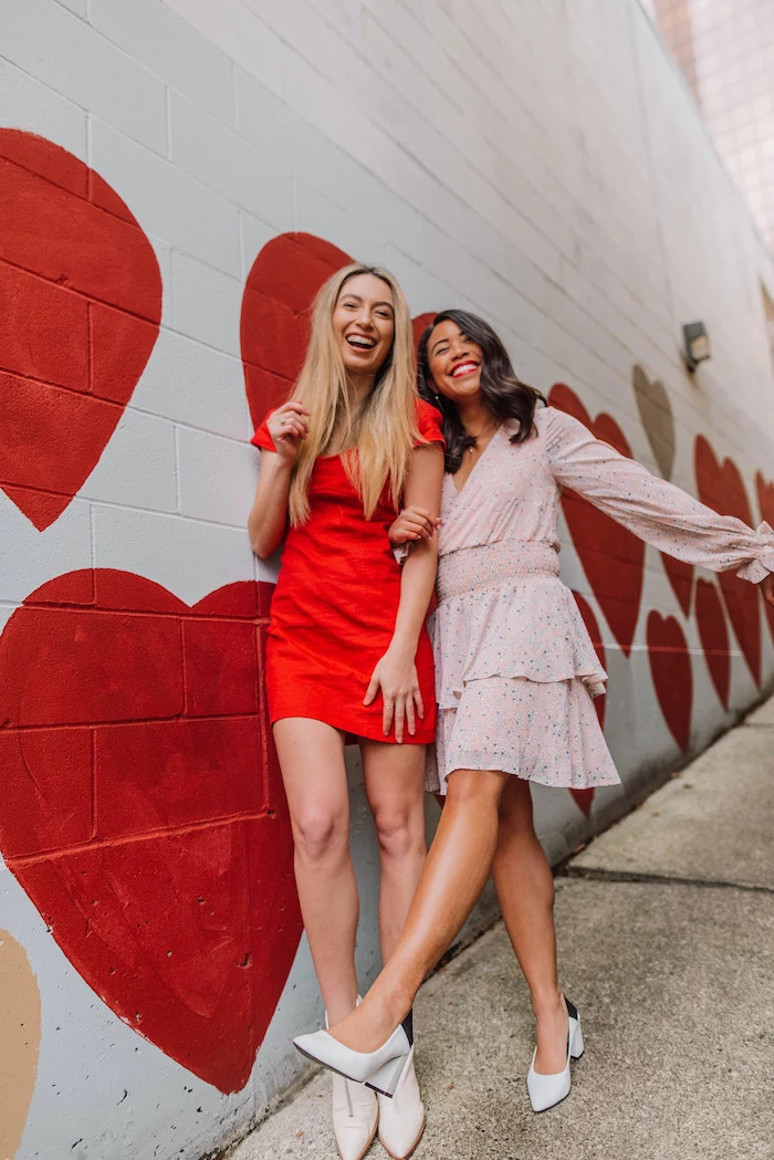 two women laughing, leaning on white wall, hearts drawn on it, valentines day dresses, wearing red and pink dresses