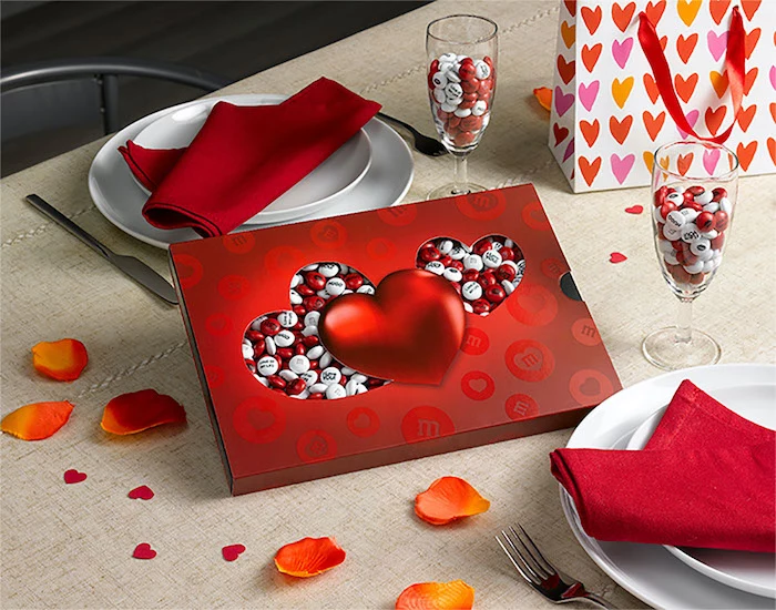 box of red and white m and ms, valentines gifts for her, placed on a table with dinner plates, red napkins in the plates