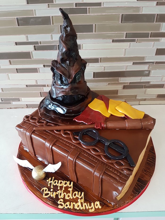 one tier cake in the shape of a book, happee birthdae, sorting hat and wand, gryffindor scarf and glasses on top