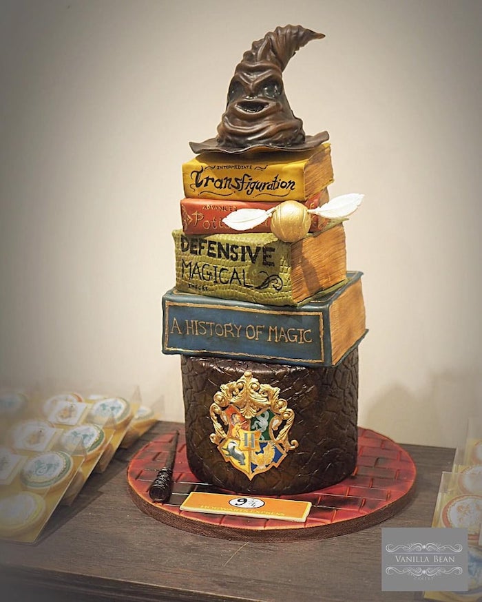 happy birthday harry cake, layers of cake in the shape of books, sorting hat made of fondant on top, placed on wooden table