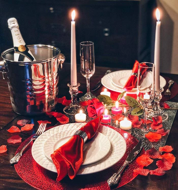dinner table set for two, red napkins and rose petals, candles and candlesticks on it, valentine's day decoration ideas