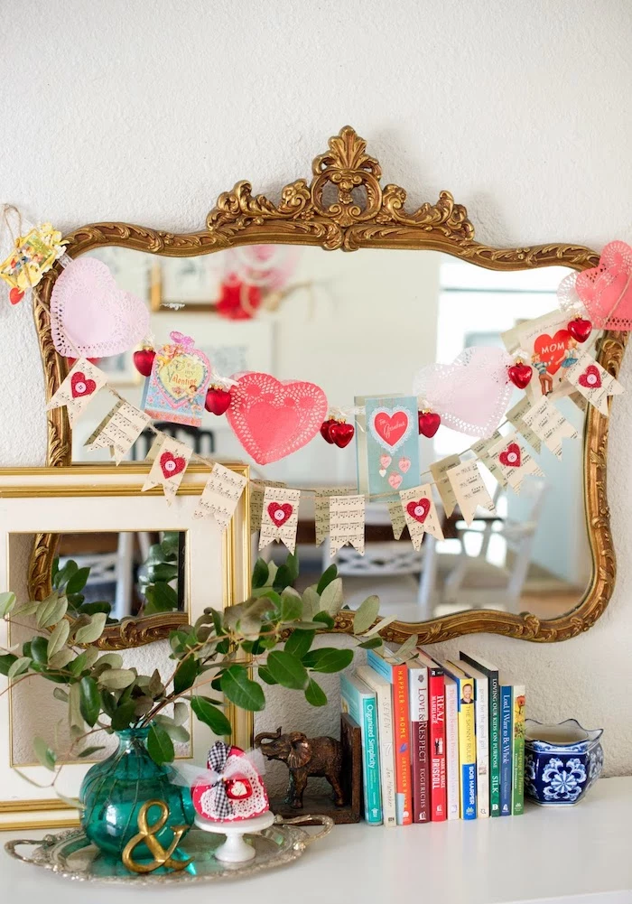 heart garlands hanging over vintage mirror, valentines day decor ideas, books and cases arranged on white surface
