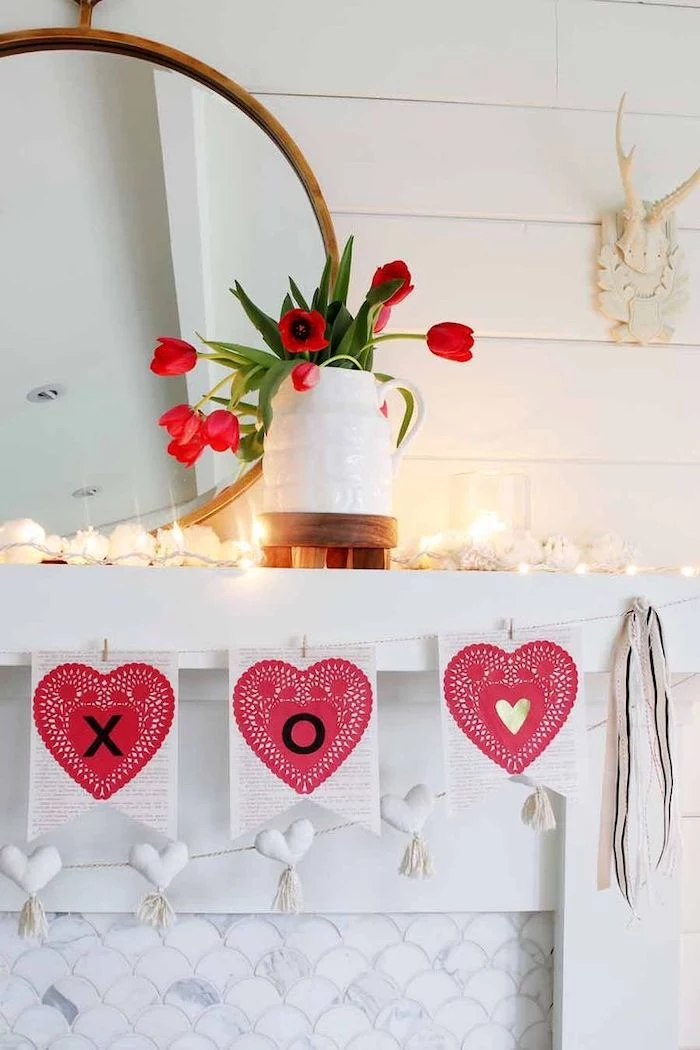 heart garland hanging over white mantel, valentines day decor ideas, lots of candles on top, bouquet of red tulips