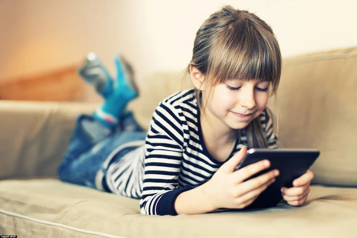 little girl lying on a sofa, holding a tablet, playing games, children's games, wearing jeans and black and white striped blouse