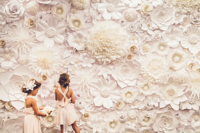 two girls wearing white dresses, standing in front of a large backdrop, covered with white paper flowers, giant paper flowers