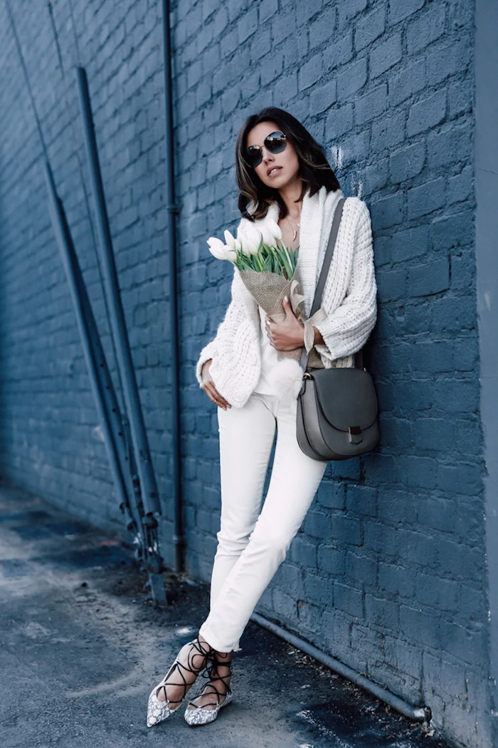woman leaning on black brick wall, red valentines day dress, wearing white pants and cardigan, holding bouquet of tulips