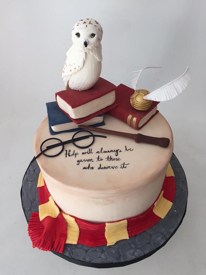 one tier ckae with white fondant, hagrid cake, red and yellow gryffindor scarf made of fondant around it, hedwig on top