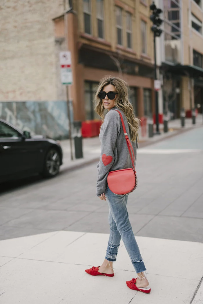 woman walking on sidewalk, wearing grey sweater and jeans, red shoes and bag, red valentines day dress