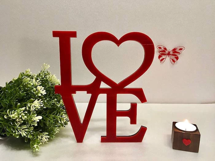 red wooden love sign, valentine lights, small candle and green bouquet next to it, placed on white surface