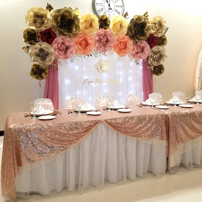 large table covered with rose gold sequinned cloth, diy tissue paper flowers, white tulle with fairy lights, large paper flowers on top