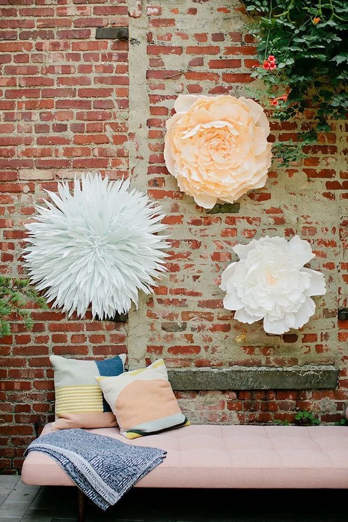 three large paper flowers, hanging on a brick wall, over a pink sofa with throw pillows, how to make paper flowers