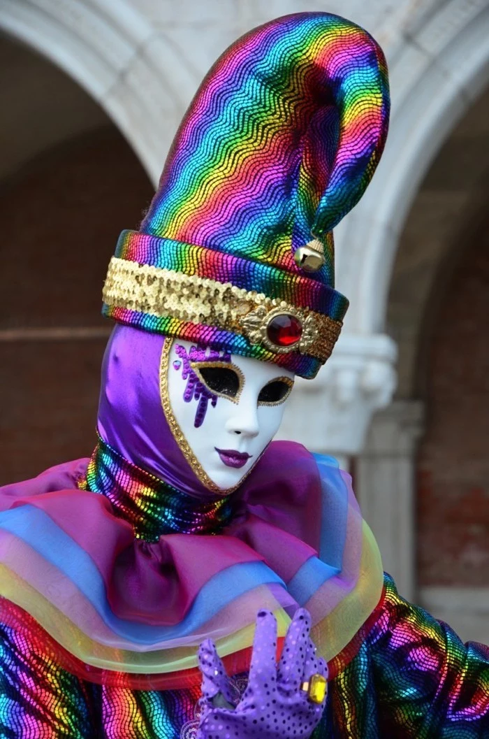 man wearing a full costume, dressed in rainbow colors, wearing a mask and large hat, mardi gras mask