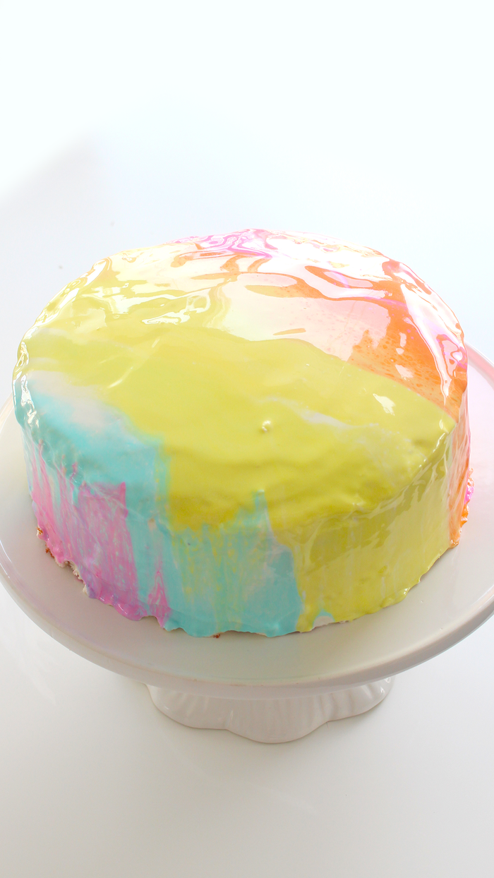 one tier cake with colorful glaze on top, pink yellow blue and orange, placed on white cake stand, mirror glaze