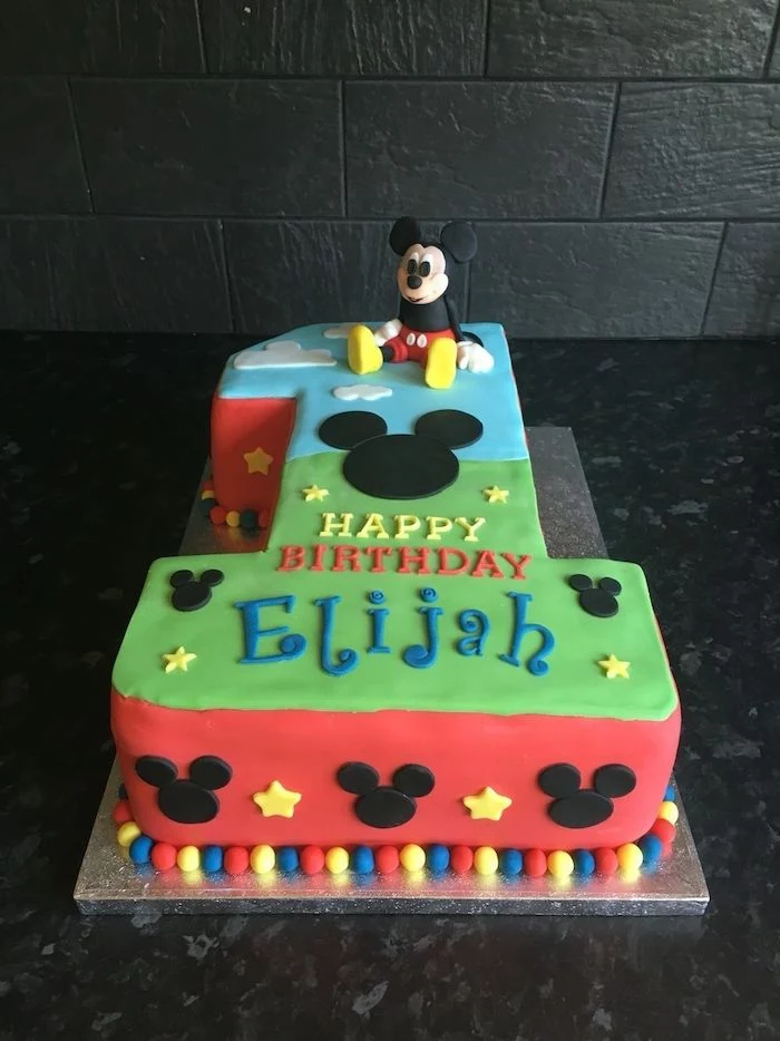 cake in the shape of the number one, mickey mouse 1st birthday cake, covered with blue green and red fondant