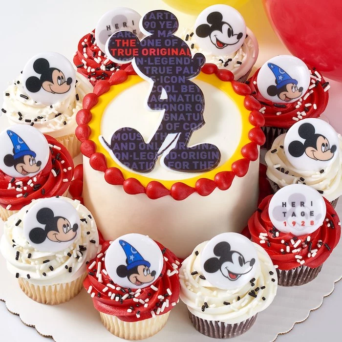 one tier cake, covered with white fondant, mickey mouse 1st birthday cake, cupcakes arranged around it