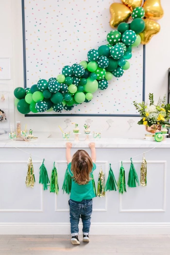 green and gold balloon hanging on white wall, st patricks day wreath, over a desserts table, green and gold tassel garland