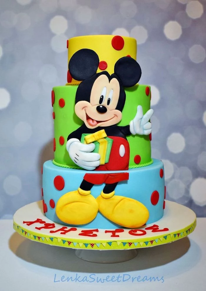 three tier cake, covered with yellow green and blue fondant, mickey mouse cake ideas, placed on white cake stand