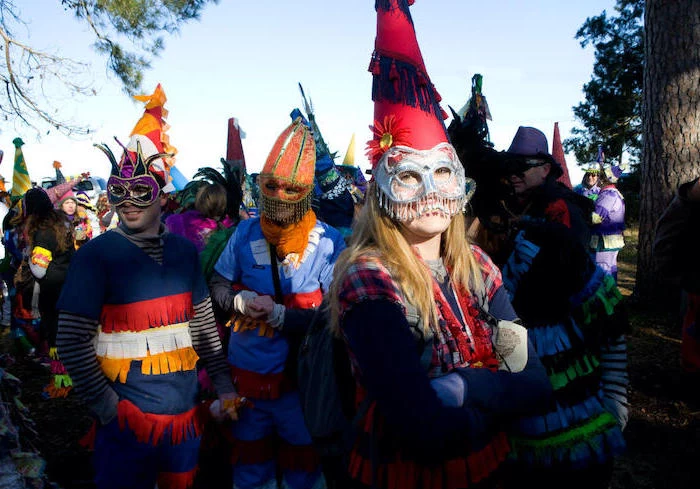 men and women gathered outside, wearing different costumes, male masquerade masks, large hats and masks