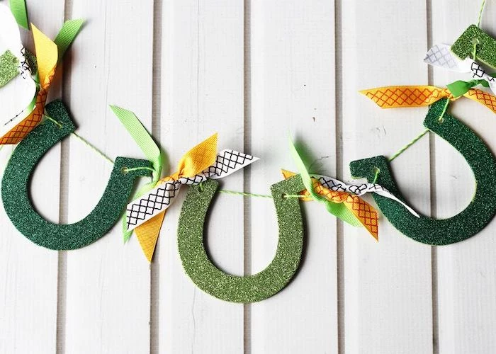 step by step diy tutorial, horseshoe garland made with green glitter, st patrick's day crafts, hanging on white wooden wall