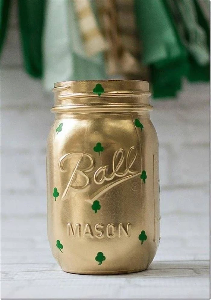 mason jar painted with gold, small green shamrocks painted on it, st patrick's day wreath, placed on white surface