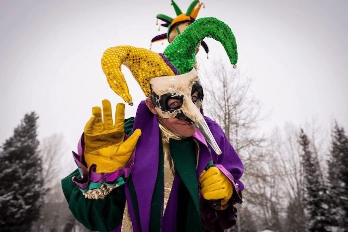 man wearing a purple green and gold costume, jester hat, masquerade masks for women, white mask with long nose
