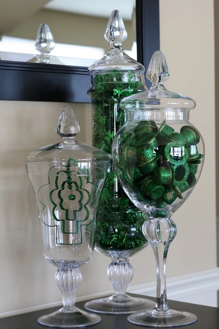 tall glass candy jars, filled with green glitter hats and garlands with shamrocks, st patricks day crafts, placed on black surface