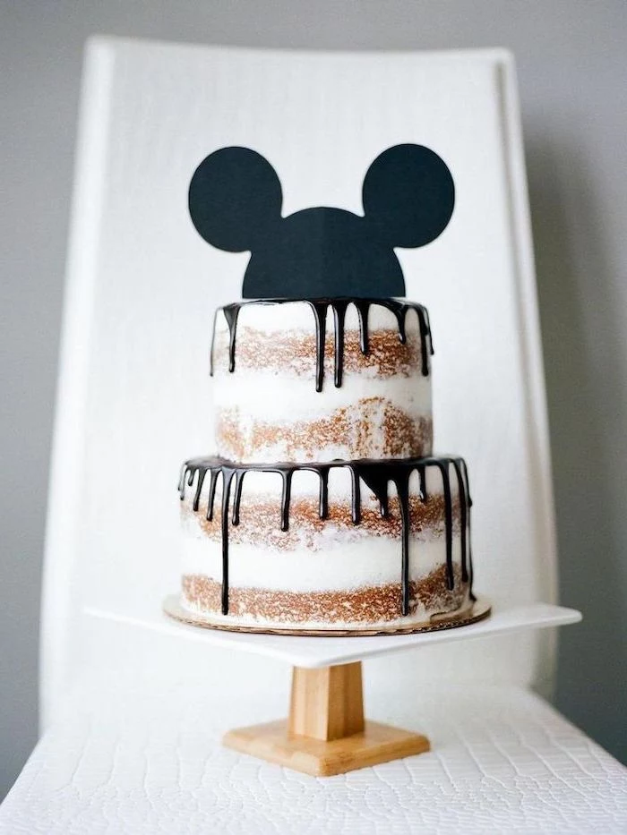 two tier cake, chocolate dripping from the sides, mickey mouse cake decorations, placed on white wooden cake stand