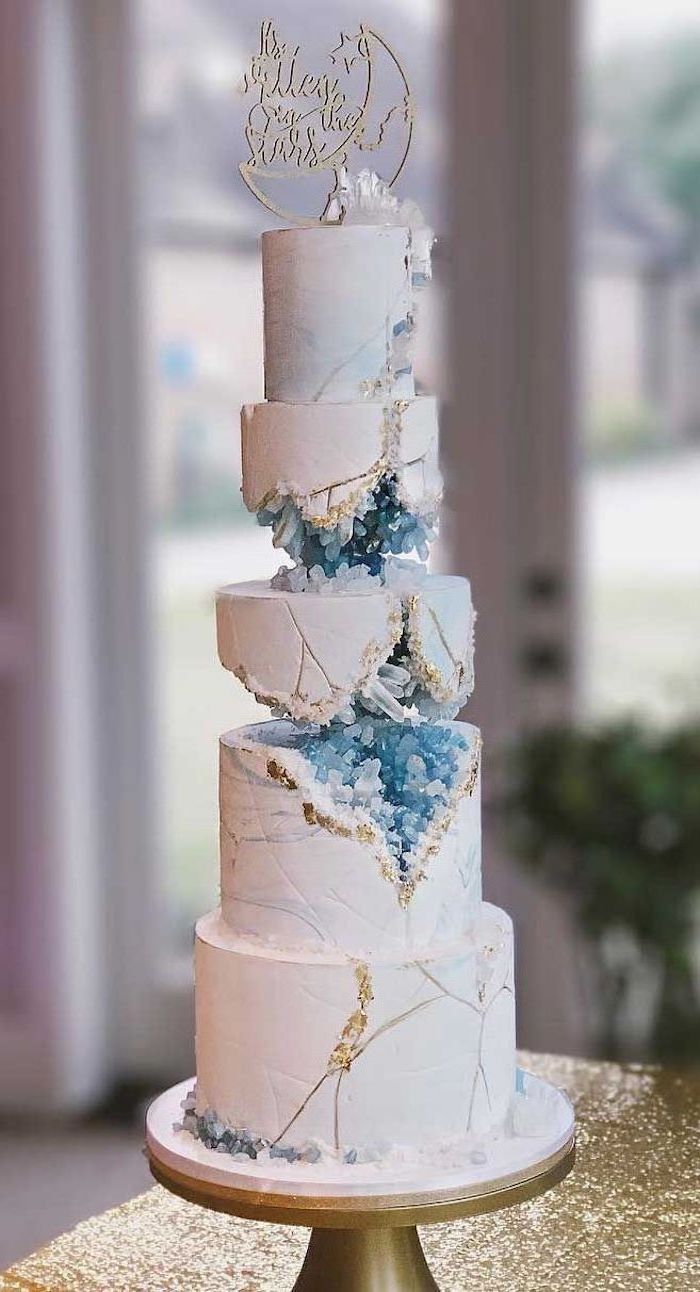 five tier cake, covered with white fondant, decorated with blue rock candy, what is a geode cake, placed on gold cake stand