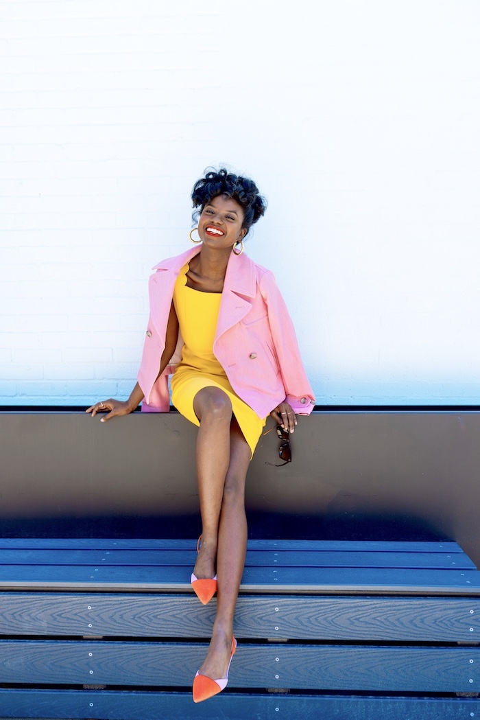woman with black hair sitting on a bench, wearing yellow drress, easter outfits, pink blazer and flats