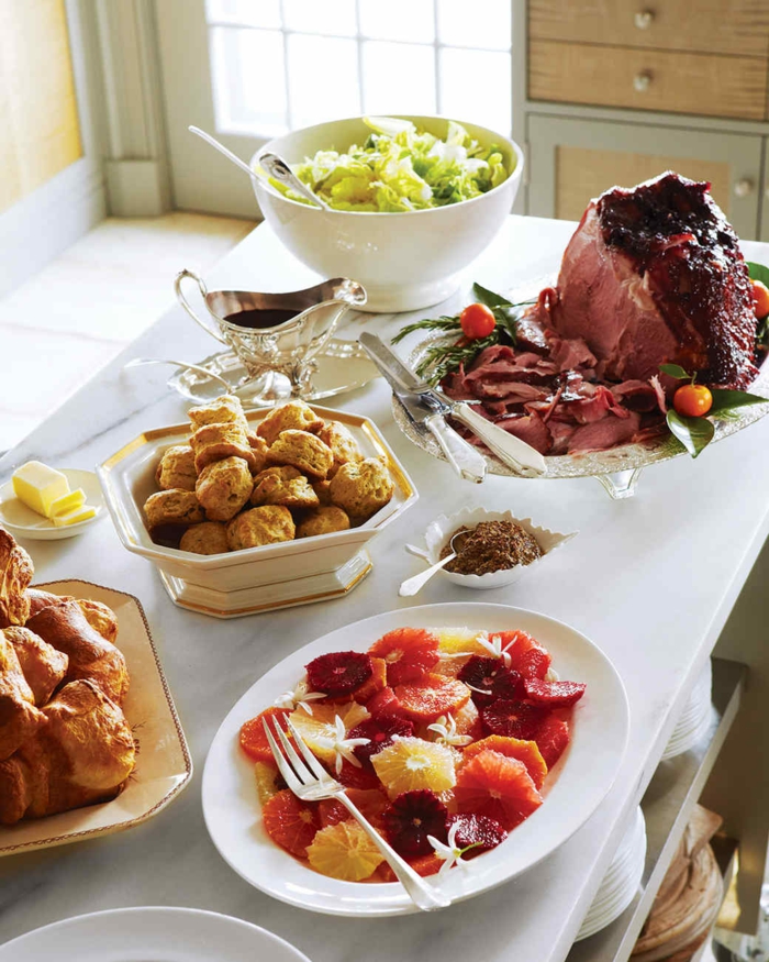 white table filled with plates and bowls, large piece of ham and bread, salad and fruits, easter lunch ideas