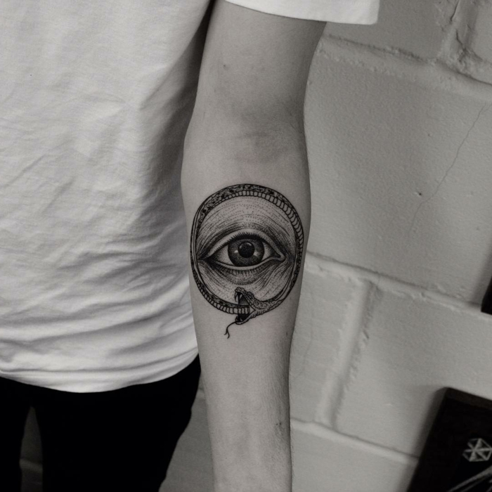 forearm tattoo, eye surrounded by a snake, ouroboros tattoo, black and white photo, white brick wall background