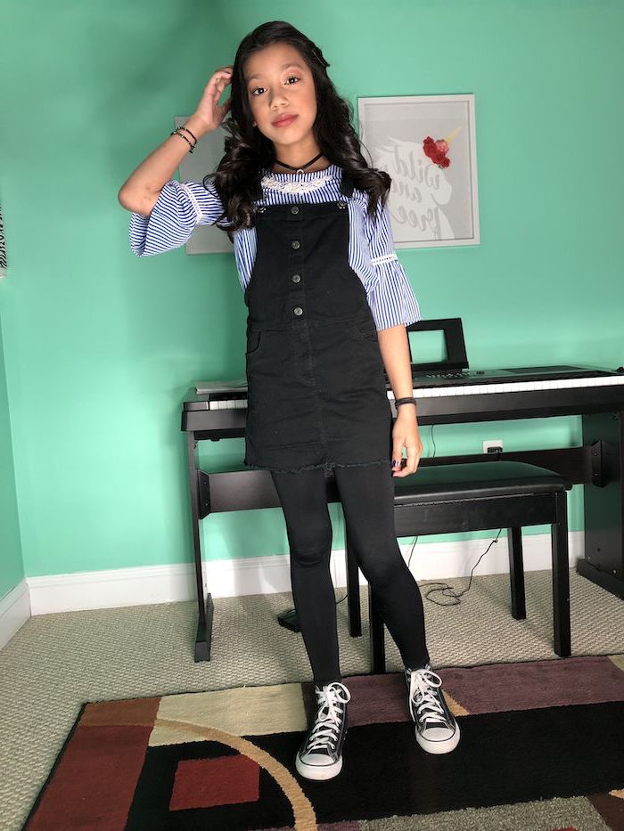 girl wearing black denim overalls, blue and white striped shirt, fall outfits for girls, black low top converse shoes